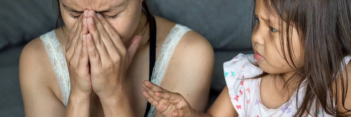 Eine Frau mit Symptomen von Stress sitzt neben ihrer Tochter auf dem Sofa und hält sich die Hände vors Gesicht.