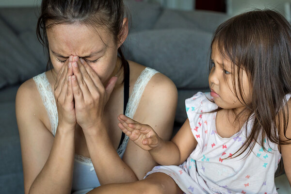Eine Frau mit Symptomen von Stress sitzt neben ihrer Tochter auf dem Sofa und hält sich die Hände vors Gesicht.