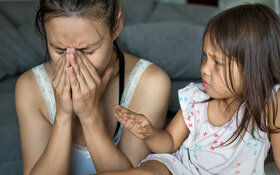 Eine Frau mit Symptomen von Stress sitzt neben ihrer Tochter auf dem Sofa und hält sich die Hände vors Gesicht.