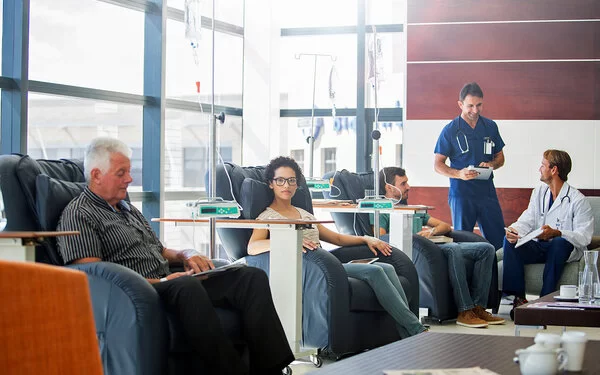 Mehrere Patienten und Patientinnen sitzen in einem Raum und erhalten eine ambulante Chemotherapie, sie werden dabei von zwei Ärzten betreut.