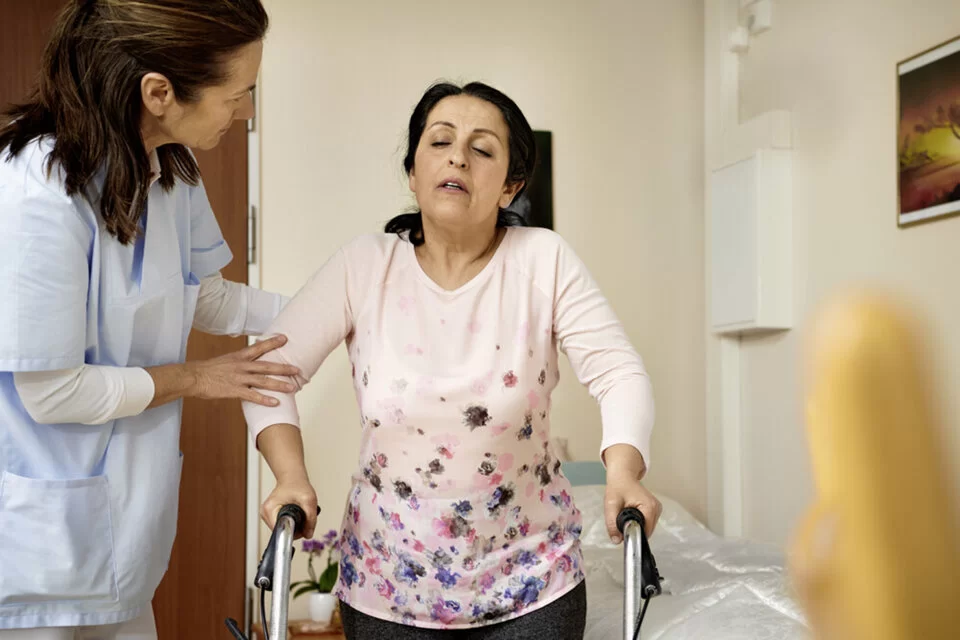 Eine Frau hält sich am Rollator fest, begleitet von einer Pflegefachperson. In der ambulanten Pflege kann ein Pflegedienst bei der Bewegung helfen.