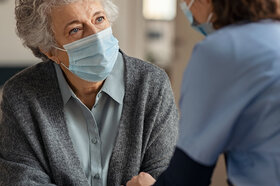 Eine Pflegerin versorgt eine ältere Frau. Beide tragen eine medizinische Maske. Auch pflegende Angehörige brauchen Schutz.