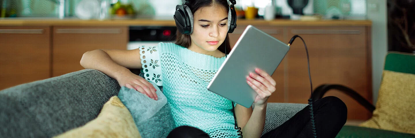 Mädchen mit Kopfhörern sitzt auf dem Sofa und schaut auf ein Tablet.