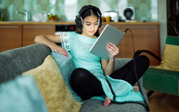 Mädchen mit Kopfhörern sitzt auf dem Sofa und schaut auf ein Tablet.