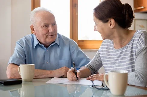 Ein älterer Mann und eine jüngere Frau sitzen zusammen am Küchentisch. Sie reden miteinander. Die Frau macht sich Notizen auf einem Blatt, das vor ihr liegt. Auf dem Tisch stehen zwei Kaffeetassen, im Hintergrund ist ein Fenster zu sehen.