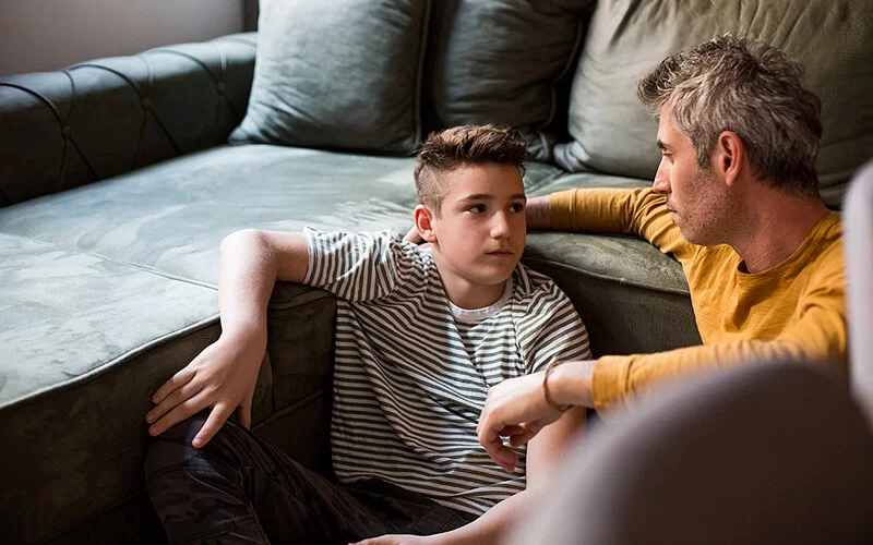 Ein Mann sitzt neben einem Jungen. Beide lehnen sich an ein Sofa an.