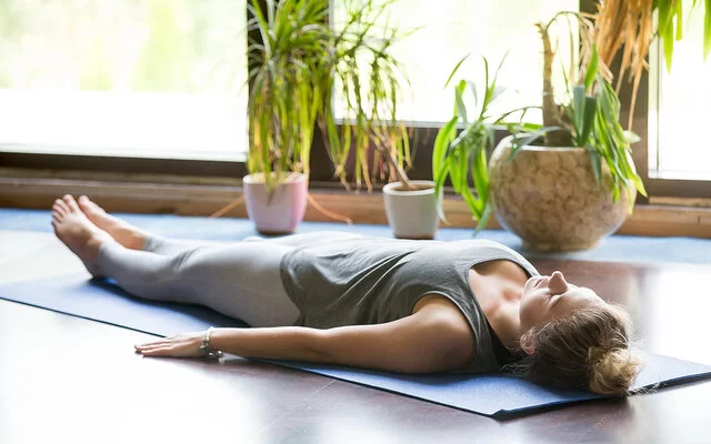 Eine Frau in Sportkleidung liegt mit dem Rücken auf einer Yogamatte und hat die Augen geschlossen. Im Hintergrund zu sehen sind Topfpflanzen und bodentiefe Fenster.