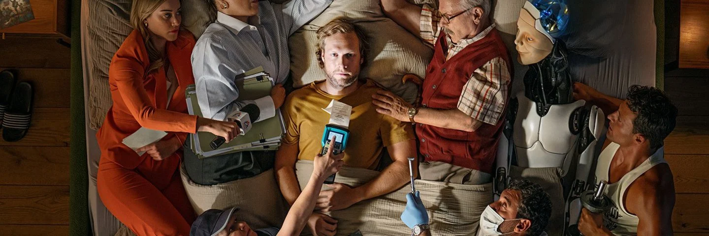 Ein Mann liegt auf einem Bett, umgeben von Menschen in verschiedenen Berufen und einem Roboter, die alle auf ihn zeigen oder ihn berühren.