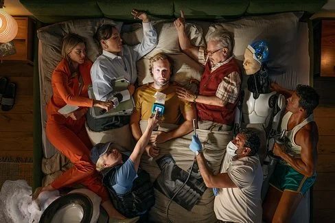 Ein Mann liegt auf einem Bett, umgeben von Menschen in verschiedenen Berufen und einem Roboter, die alle auf ihn zeigen oder ihn berühren.