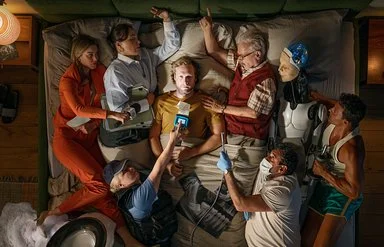Ein Mann liegt auf einem Bett, umgeben von Menschen in verschiedenen Berufen und einem Roboter, die alle auf ihn zeigen oder ihn berühren.