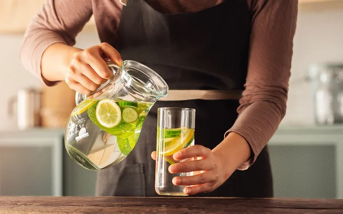 Eine Frau schenkt sich aus einer Karaffe Wasser in ein Glas ein. Das Wasser enthält einige Zitronen- und Gurkenscheiben.