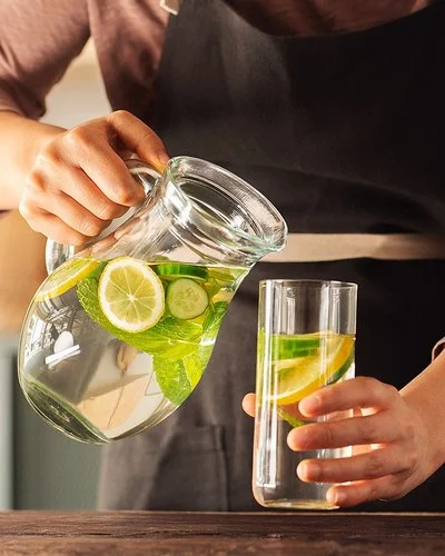 Eine Frau schenkt sich aus einer Karaffe Wasser in ein Glas ein. Das Wasser enthält einige Zitronen- und Gurkenscheiben.