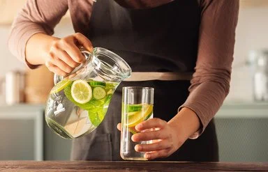 Eine Frau schenkt sich aus einer Karaffe Wasser in ein Glas ein. Das Wasser enthält einige Zitronen- und Gurkenscheiben.