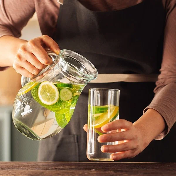 Eine Frau schenkt sich aus einer Karaffe Wasser in ein Glas ein. Das Wasser enthält einige Zitronen- und Gurkenscheiben.