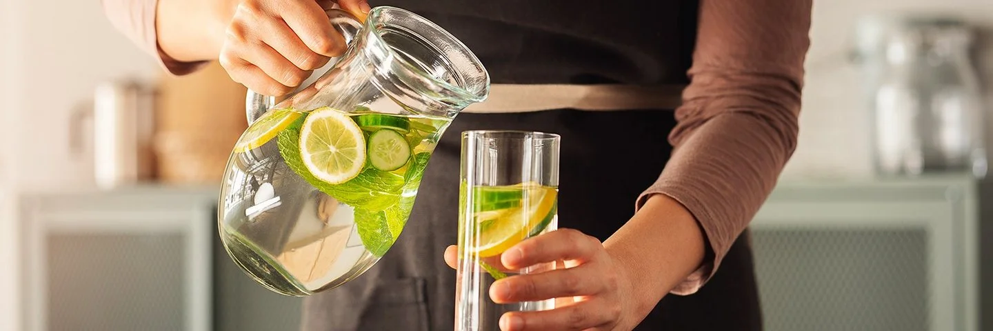 Eine Frau schenkt sich aus einer Karaffe Wasser in ein Glas ein. Das Wasser enthält einige Zitronen- und Gurkenscheiben.