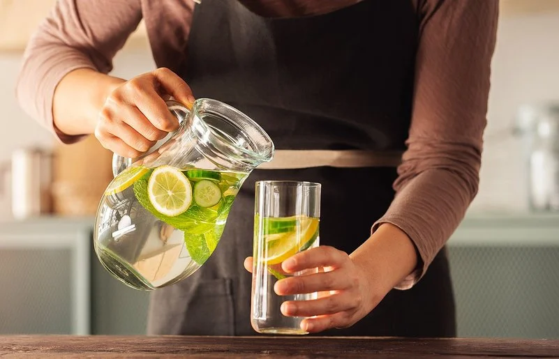 Eine Frau schenkt sich aus einer Karaffe Wasser in ein Glas ein. Das Wasser enthält einige Zitronen- und Gurkenscheiben.