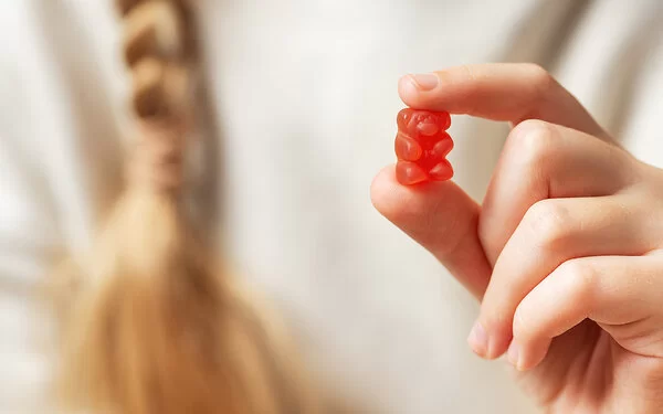 Im Vordergrund eine Kinderhand, die ein rotes Gummibärchen zwischen Zeigefinger und Daumen hält. Im Hintergrund in der Unschärfe der weiße Pullover und der Zopf eines Mädchens.
