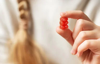 Im Vordergrund eine Kinderhand, die ein rotes Gummibärchen zwischen Zeigefinger und Daumen hält. Im Hintergrund in der Unschärfe der weiße Pullover und der Zopf eines Mädchens.