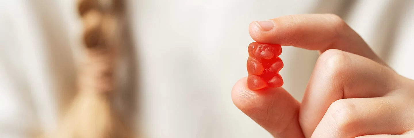 Im Vordergrund eine Kinderhand, die ein rotes Gummibärchen zwischen Zeigefinger und Daumen hält. Im Hintergrund in der Unschärfe der weiße Pullover und der Zopf eines Mädchens.