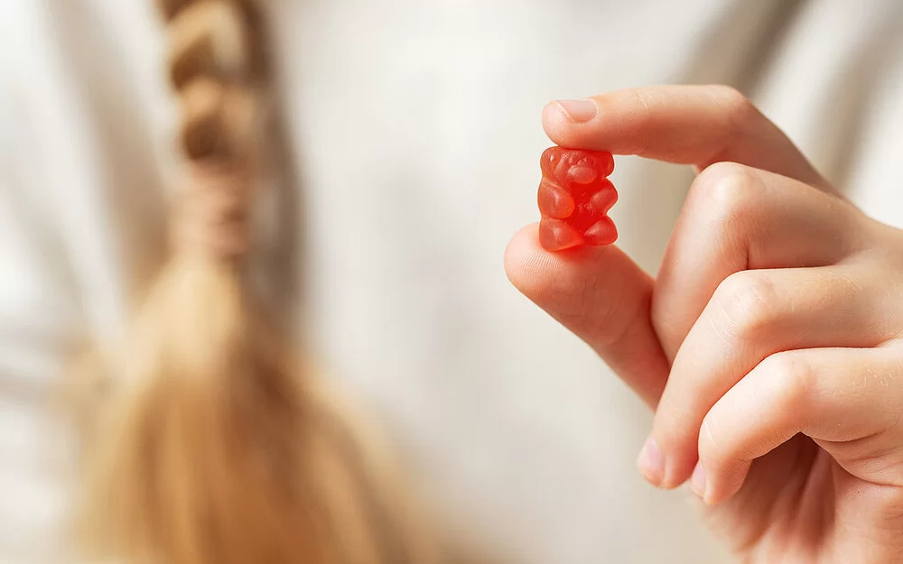 Im Vordergrund eine Kinderhand, die ein rotes Gummibärchen zwischen Zeigefinger und Daumen hält. Im Hintergrund in der Unschärfe der weiße Pullover und der Zopf eines Mädchens.