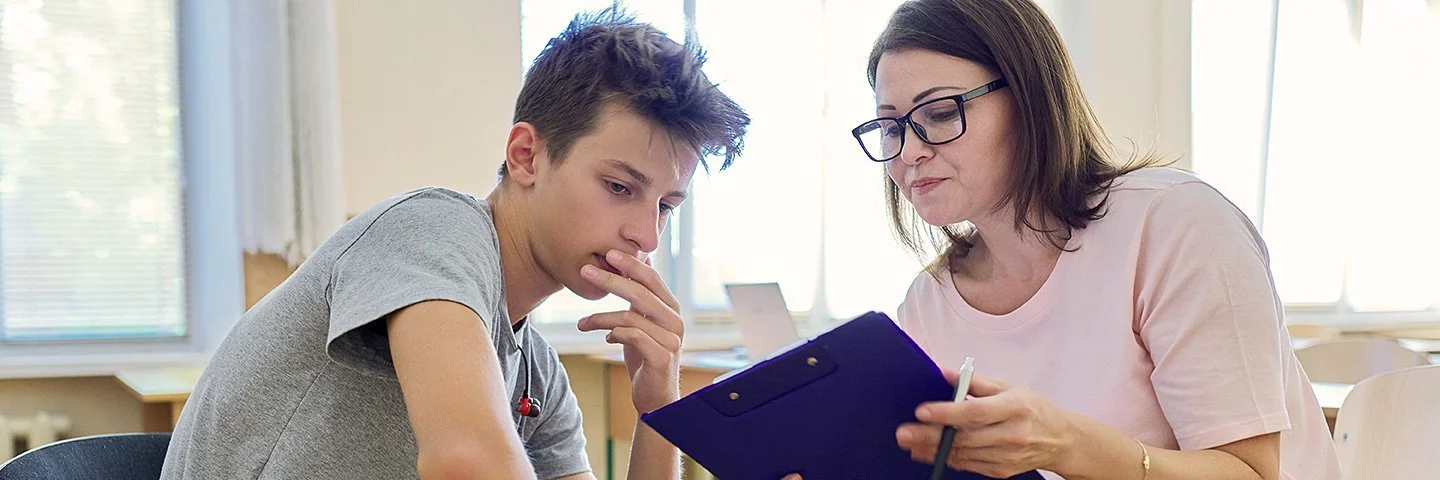 Eine Lehrerin oder Beraterin hält ein Klemmbrett und spricht in einem Klassenraum mit einem Jugendlichen.