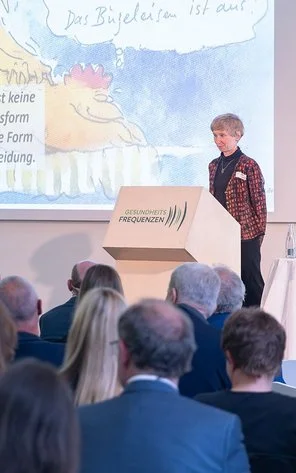 Eine Frau steht am Rednerpult neben einem Dozenten vor einer Gruppe Zuhörer.