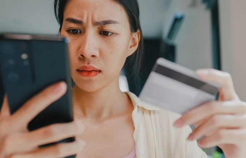 Eine junge Frau hält in der rechten Hand ein Smartphone und in der linken eine Kredit- oder andere Bankkarte. Beide befinden sich auf Augenhöhe. Sie blickt ernst auf das Display des Smartphones.