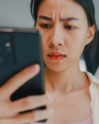 Eine junge Frau hält in der rechten Hand ein Smartphone und in der linken eine Kredit- oder andere Bankkarte. Beide befinden sich auf Augenhöhe. Sie blickt ernst auf das Display des Smartphones.