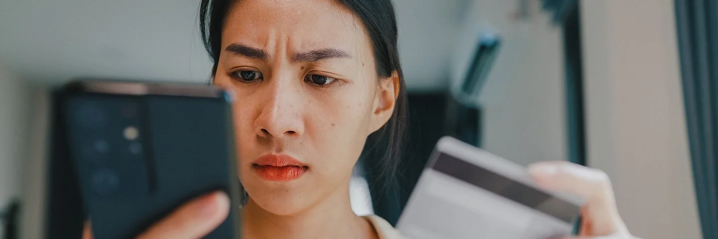 Eine junge Frau hält in der rechten Hand ein Smartphone und in der linken eine Kredit- oder andere Bankkarte. Beide befinden sich auf Augenhöhe. Sie blickt ernst auf das Display des Smartphones.