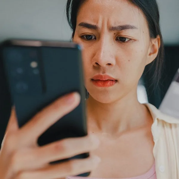 Eine junge Frau hält in der rechten Hand ein Smartphone und in der linken eine Kredit- oder andere Bankkarte. Beide befinden sich auf Augenhöhe. Sie blickt ernst auf das Display des Smartphones.