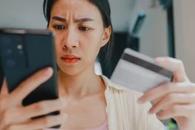 Eine junge Frau hält in der rechten Hand ein Smartphone und in der linken eine Kredit- oder andere Bankkarte. Beide befinden sich auf Augenhöhe. Sie blickt ernst auf das Display des Smartphones.