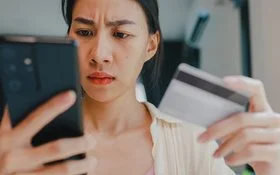 Eine junge Frau hält in der rechten Hand ein Smartphone und in der linken eine Kredit- oder andere Bankkarte. Beide befinden sich auf Augenhöhe. Sie blickt ernst auf das Display des Smartphones.