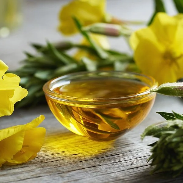 Eine kleine mit Öl gefüllte Schale steht auf einem Holztisch, daneben liegen Nachtkerzenblüten.