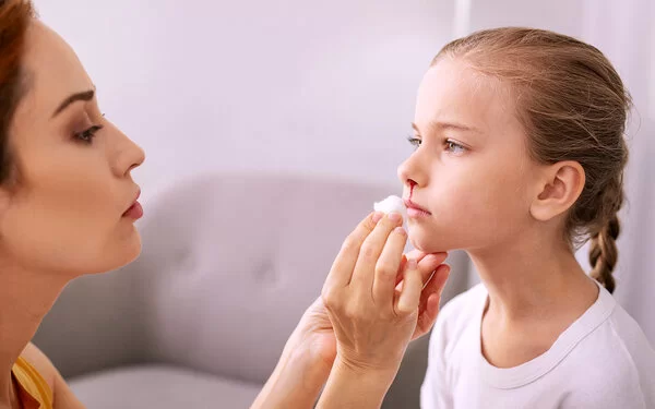 Mädchen hat Nasenblutung, ihre Mutter tupft vorsichtig das Blut weg.