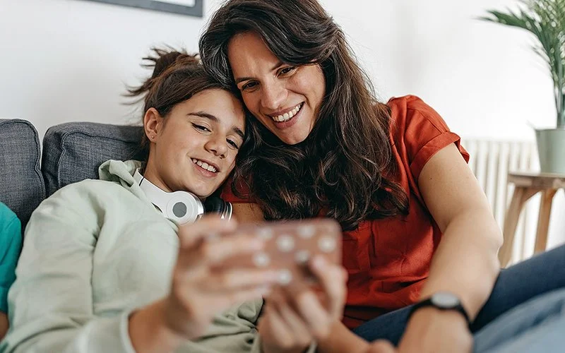 Auf einem Sofa sitzen ein Teenagermädchen und eine Frau. Das Mädchen hält ein Smartphone in der Hand. Beide blicken auf das Smartphone.