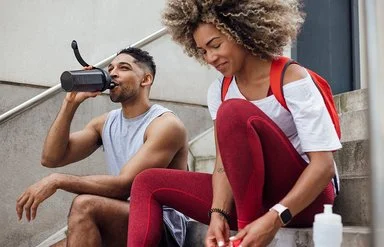 Zwei Personen in sportlicher Kleidung sitzen auf einer Treppe im Freien. Die Person links trägt ein ärmelloses graues Oberteil und trinkt aus einer schwarzen Trinkflasche. Die Person rechts trägt ein rotes Oberteil, eine rote Leggings und einen roten Rucksack. Vor ihr steht eine weiße Trinkflasche. Beide wirken entspannt und befinden sich in einer Pause.
