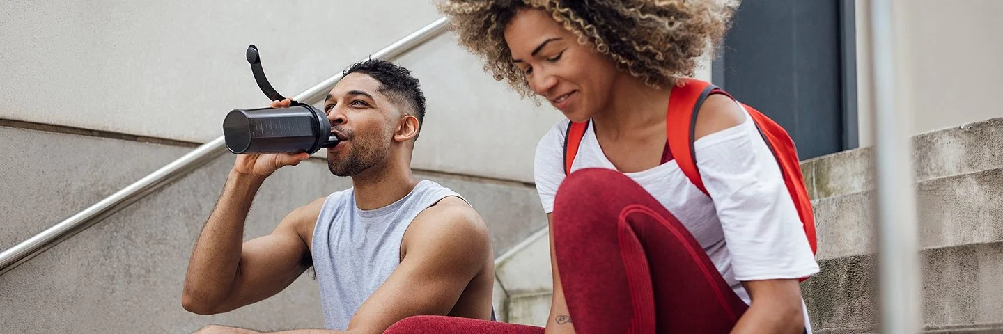 Zwei Personen in sportlicher Kleidung sitzen auf einer Treppe im Freien. Die Person links trägt ein ärmelloses graues Oberteil und trinkt aus einer schwarzen Trinkflasche. Die Person rechts trägt ein rotes Oberteil, eine rote Leggings und einen roten Rucksack. Vor ihr steht eine weiße Trinkflasche. Beide wirken entspannt und befinden sich in einer Pause.