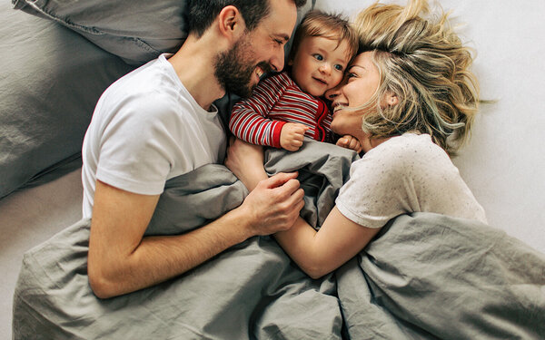 Eine junge Familie mit Baby liegt im Bett und lacht.