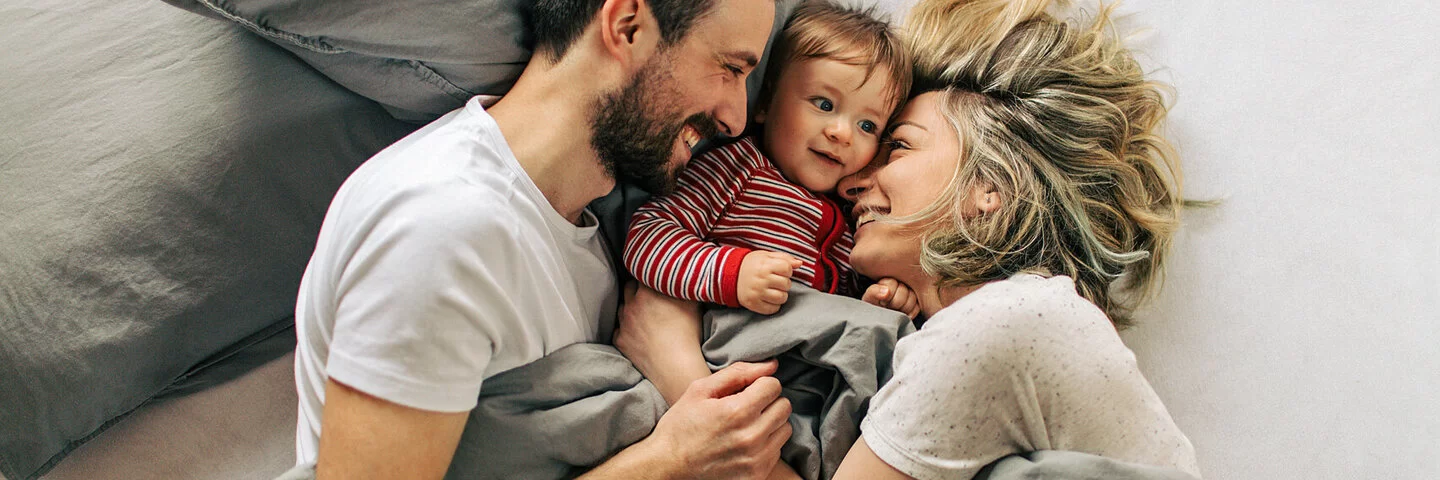 Eine junge Familie mit Baby liegt im Bett und lacht.