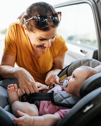 Eine Mutter schnallt lächelnd ihr Baby in Babyschale an, die rückwärtsgewandt eingebaut ist.