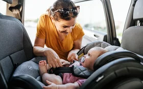 Eine Mutter schnallt lächelnd ihr Baby in Babyschale an, die rückwärtsgewandt eingebaut ist.