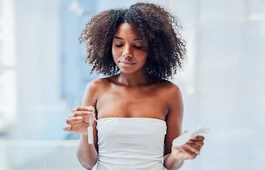 Eine junge Frau steht im Badezimmer und hält mit der einen Hand einen Tampon und mit der anderen eine Damenbinde.