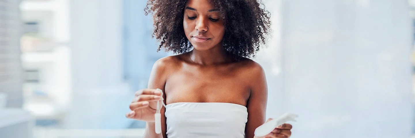 Eine junge Frau steht im Badezimmer und hält mit der einen Hand einen Tampon und mit der anderen eine Damenbinde.