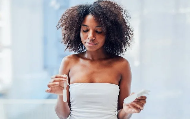 Eine junge Frau steht im Badezimmer und hält mit der einen Hand einen Tampon und mit der anderen eine Damenbinde.