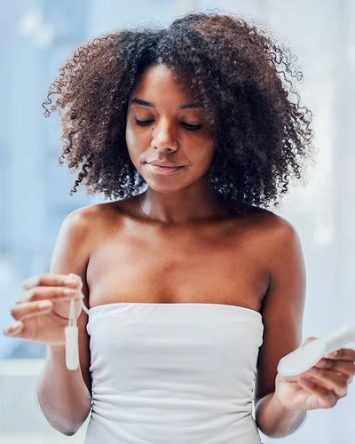 Eine junge Frau steht im Badezimmer und hält mit der einen Hand einen Tampon und mit der anderen eine Damenbinde.