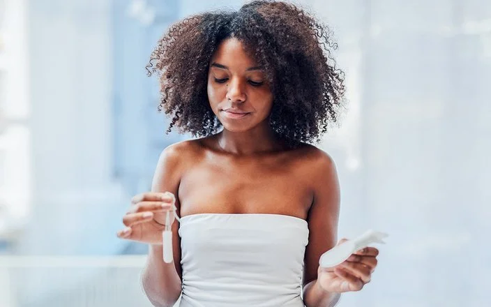 Eine junge Frau steht im Badezimmer und hält mit der einen Hand einen Tampon und mit der anderen eine Damenbinde.