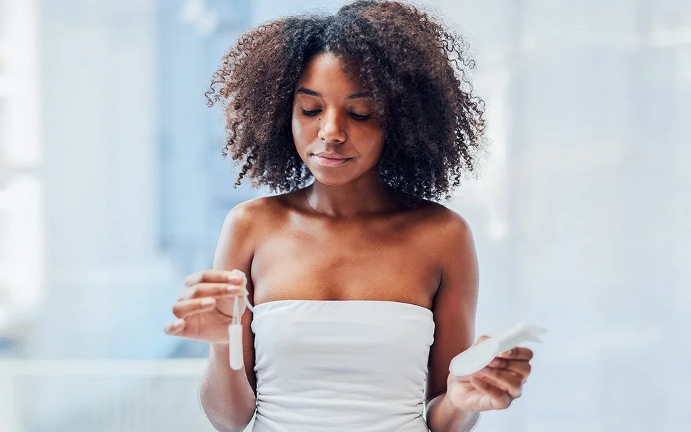 Eine junge Frau steht im Badezimmer und hält mit der einen Hand einen Tampon und mit der anderen eine Damenbinde.