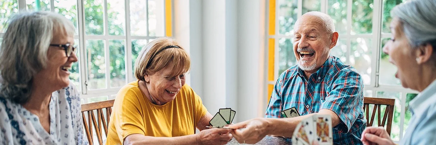 Pflege-WG: Ein älterer Mann und drei ältere Frauen sitzen zusammen an einem Tisch und spielen ausgelassen Karten.