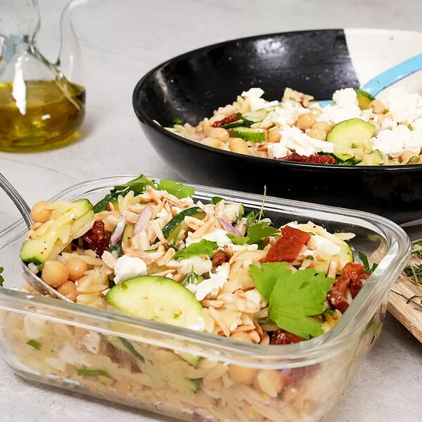 Im Vordergrund steht in einer Glasbox Orzo-Salat mit Zucchini und Kichererbsen mit Honig-Senf-Dressing nach einem Rezept von Felicitas Then. Im Hintergrund ist der Orzo-Salat auf einem schwarz-weißen Teller angerichtet. Rechts liegt eine halbe Zitrone, hinten links steht ein Ölkännchen.