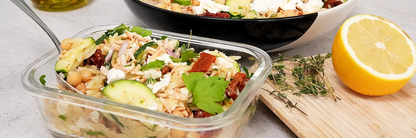 Im Vordergrund steht in einer Glasbox Orzo-Salat mit Zucchini und Kichererbsen mit Honig-Senf-Dressing nach einem Rezept von Felicitas Then. Im Hintergrund ist der Orzo-Salat auf einem schwarz-weißen Teller angerichtet. Rechts liegt eine halbe Zitrone, hinten links steht ein Ölkännchen.